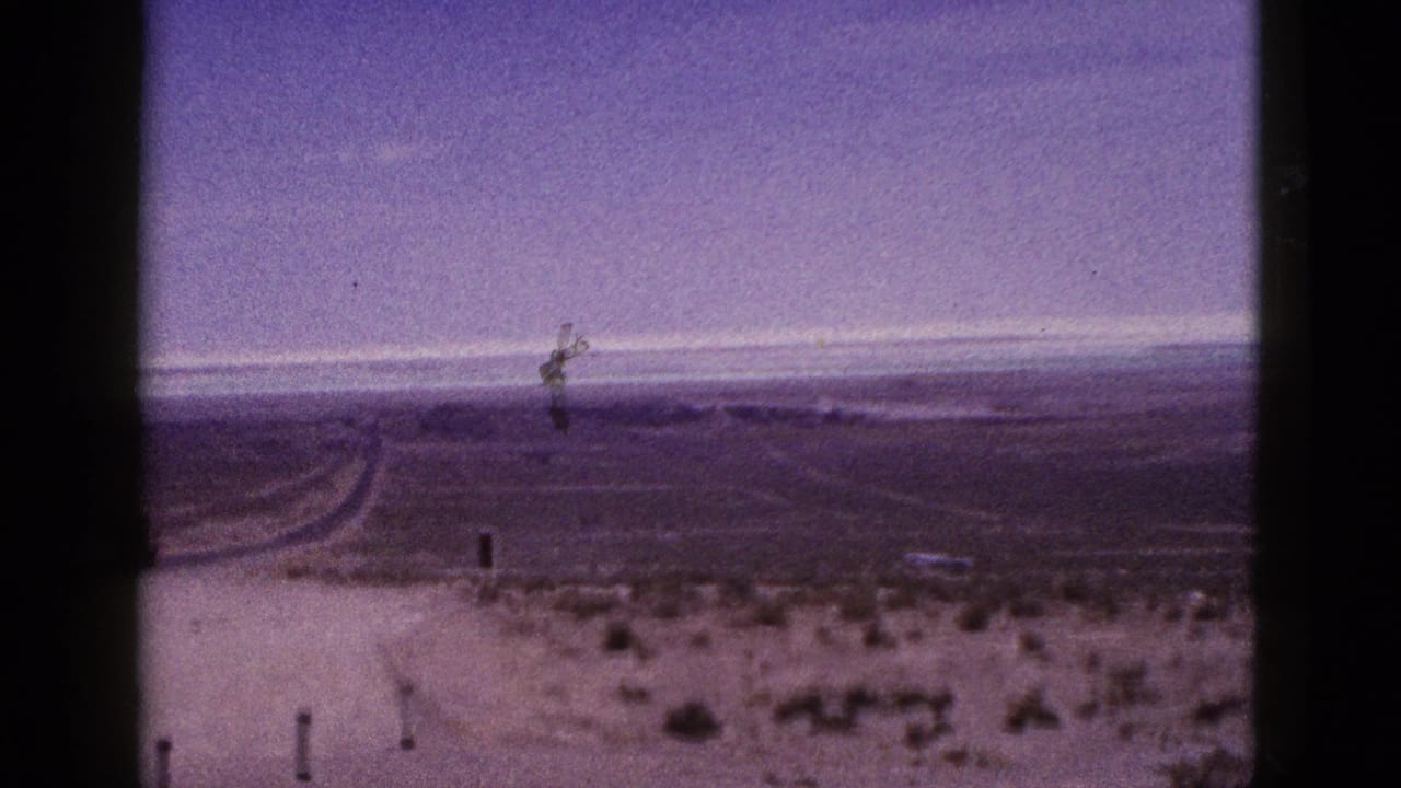 1953 California 8mm frame showing a sparse desert road curving away through flat terrain with shrubs, posts, and dark film borders.