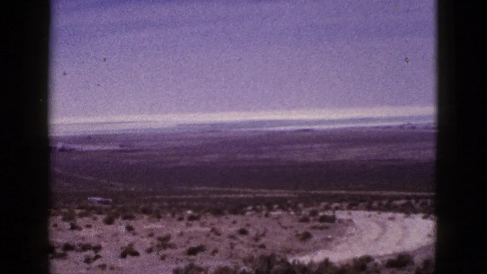 1953 California home-movie frame showing a wide desert vista with scrub vegetation, pale sand, distant flat land, and 8mm border marks.