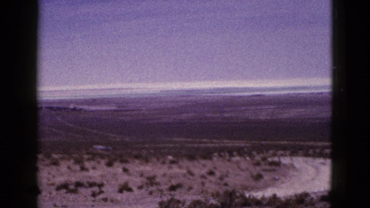 1953 California 8mm frame showing open desert scrub, a distant road corridor, low hills, hazy sky, and visible film-strip borders.