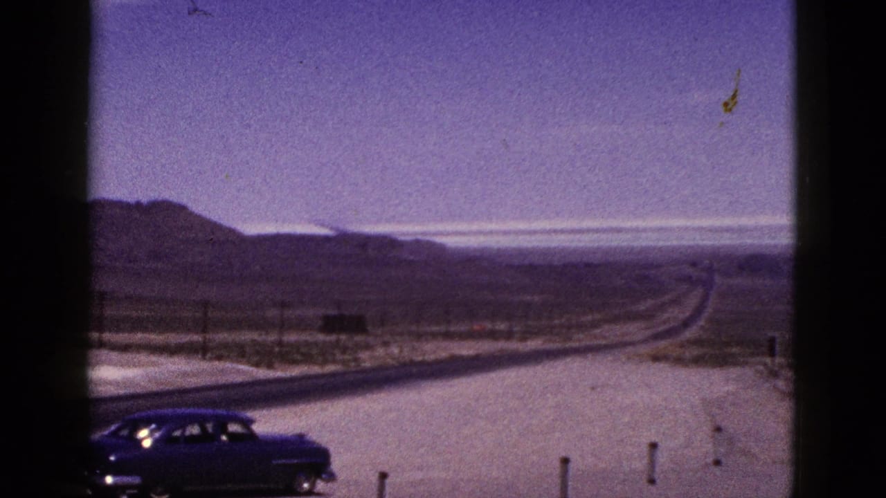 Dark vintage sedan beside a desert highway with low hills and utility poles in the distance.