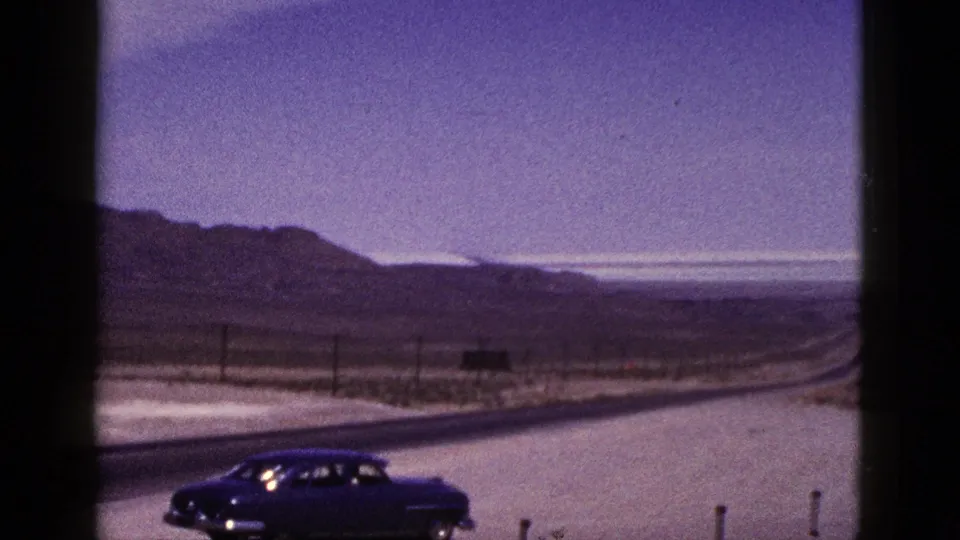 1953 California 8mm frame with a dark vintage sedan in the foreground, a curving two-lane desert highway, low hills, poles, and road markers.