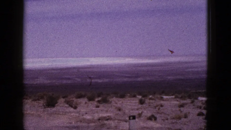 1953 California desert landscape on 8mm, showing scrubby roadside ground, a broad open plain, distant hills, hazy sky, and film-edge borders.