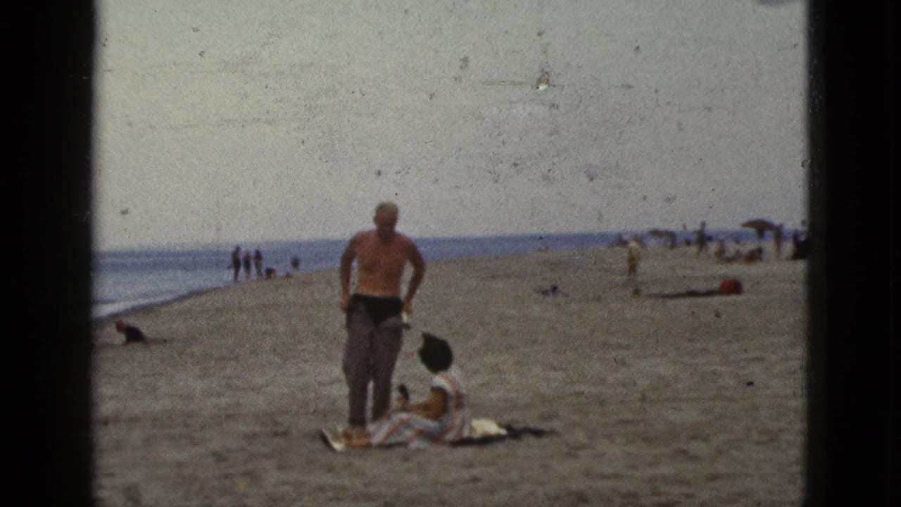 Man stands on a sandy beach near a seated woman on a blanket, with ocean and distant beachgoers behind them.