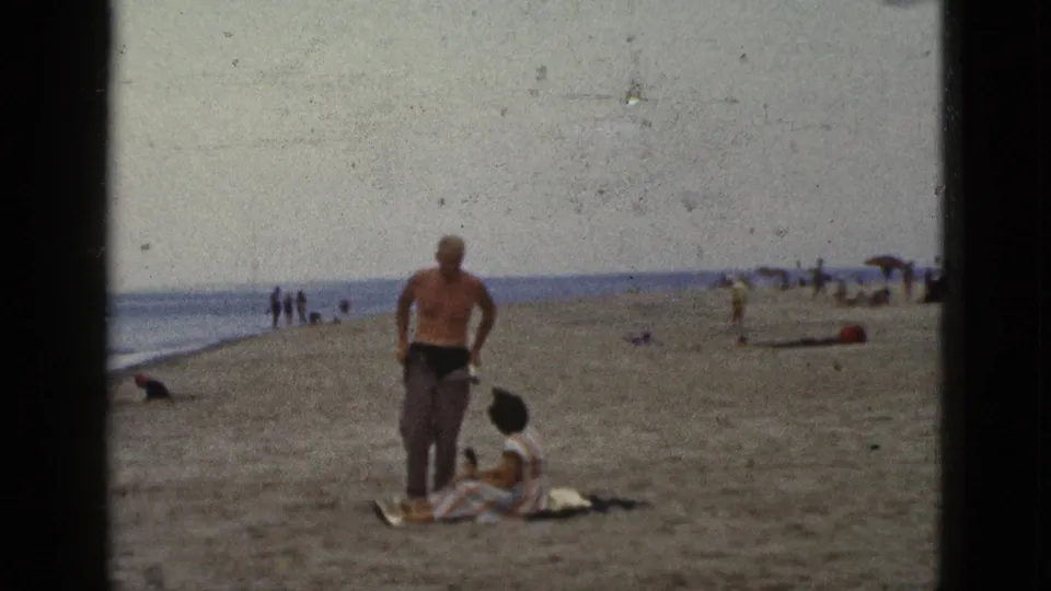 1953 California 8mm frame showing a man beside a seated woman on a beach blanket, with surf, umbrellas, and distant beachgoers in the background.