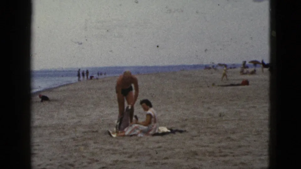 1953 California beach scene with a man bending near clothing or a towel beside a seated woman, while distant people and umbrellas line the sand.