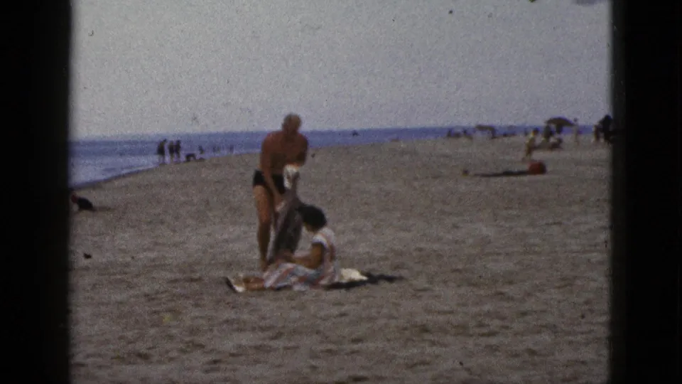 1953 California 8mm frame of a man adjusting clothing near a seated woman on a beach blanket, with surf and small groups in the background.