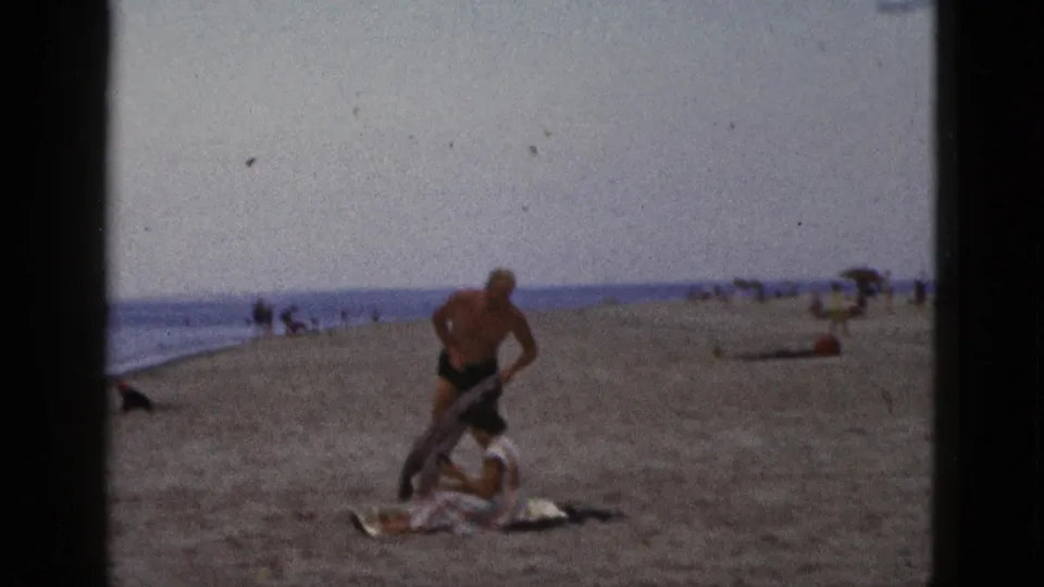 1953 California beach frame showing a man changing into swimwear beside a seated woman, with an open sandy beach and ocean horizon behind them.
