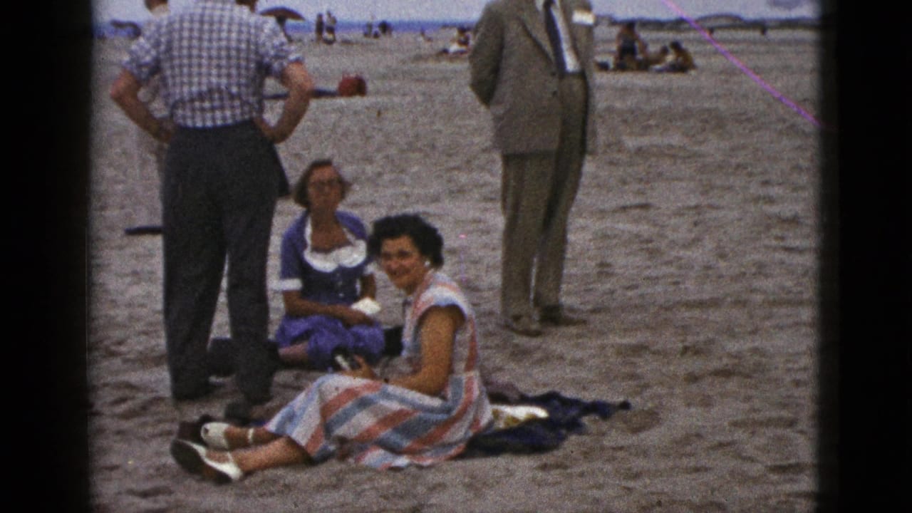 1953 California beach home-movie frame with two seated women on a blanket and several adult men standing around them.