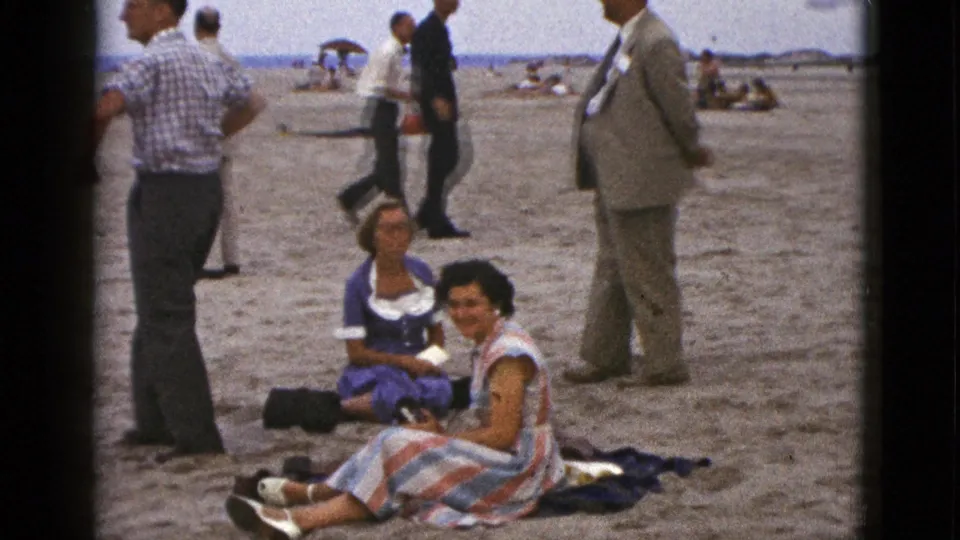 1953 California beach scene with seated women looking toward the camera, men nearby, and distant umbrellas along the shoreline.