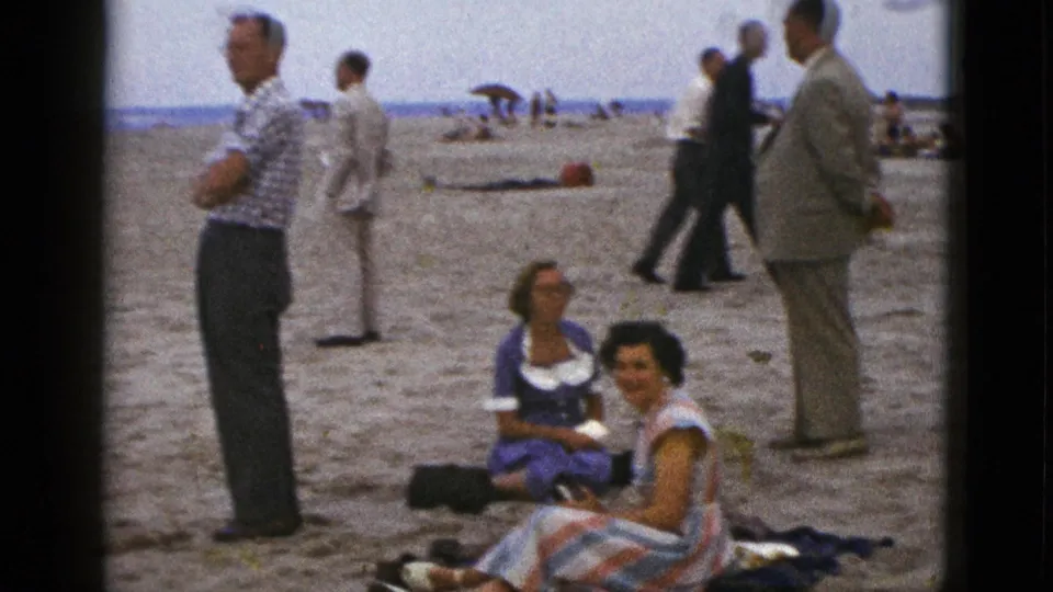 1953 California beach frame showing women seated on a blanket with men standing nearby and distant visitors along the ocean horizon.
