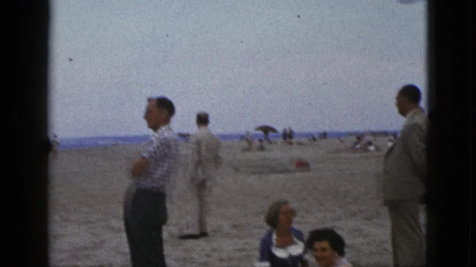 Wide 1953 California beach home-movie frame with seated women low in the shot, adult men standing, and distant beach umbrellas near the shoreline.