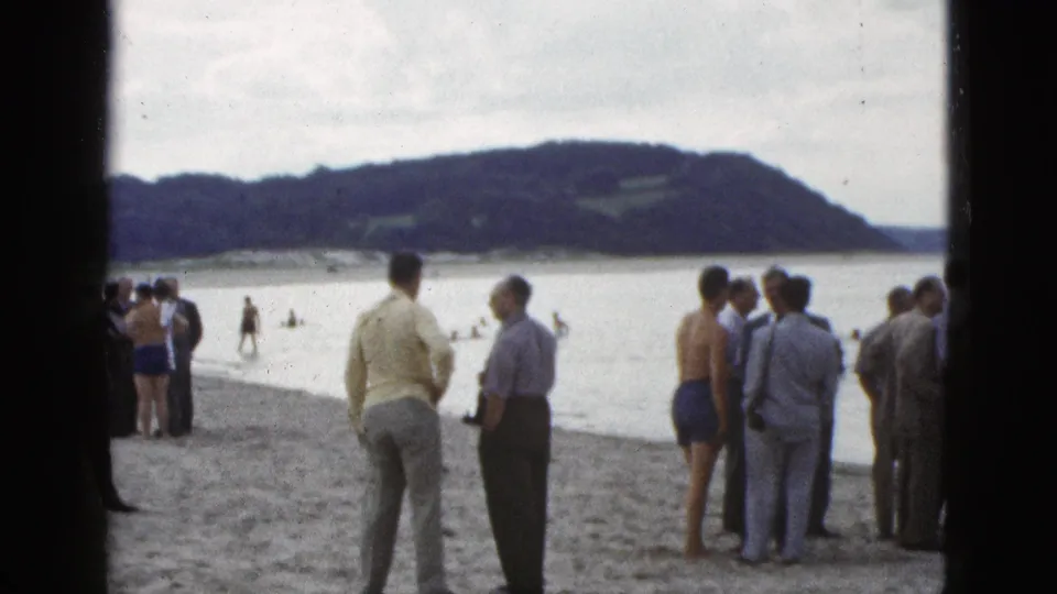 1953 California 8mm frame of suited men and beachgoers gathered on a sandy bayside shore as swimmers move offshore.