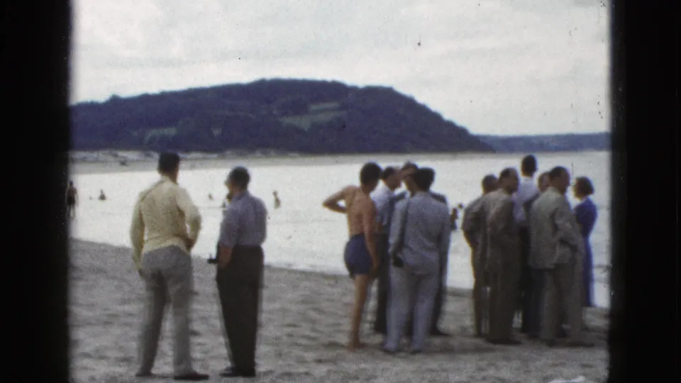 A bayside crowd in 1953 California watches the water, with men in light suits and shirts beside a shirtless swimmer on shore.