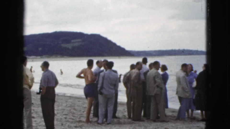1953 California home-movie frame showing a larger cluster of spectators on sand, with swimmers in the bay and a wooded hill beyond.