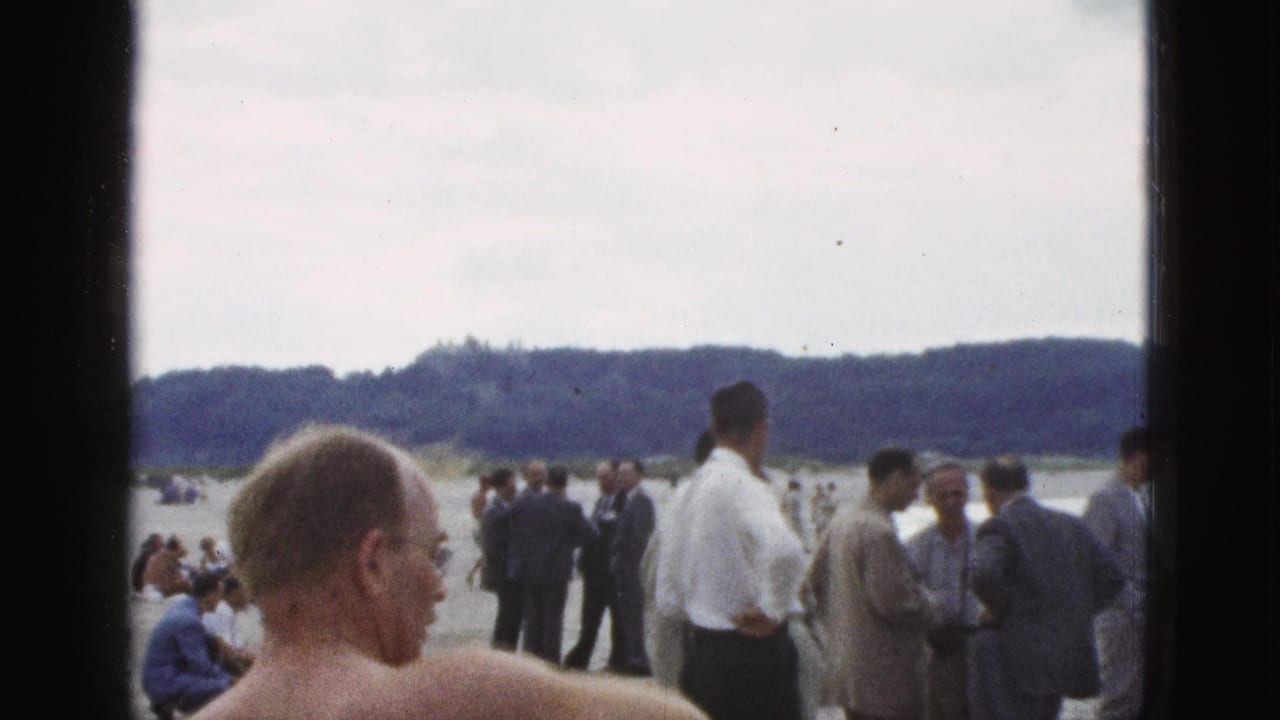 1953 California beach scene with a shirtless man in the foreground, men in suits and shirts on sand, and swimmers near a tree-lined shore.
