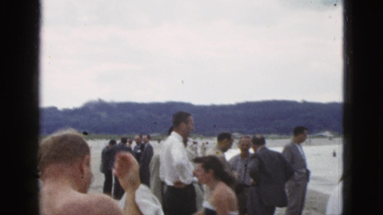 A man raises a towel near his head as beachgoers stand along the shoreline.