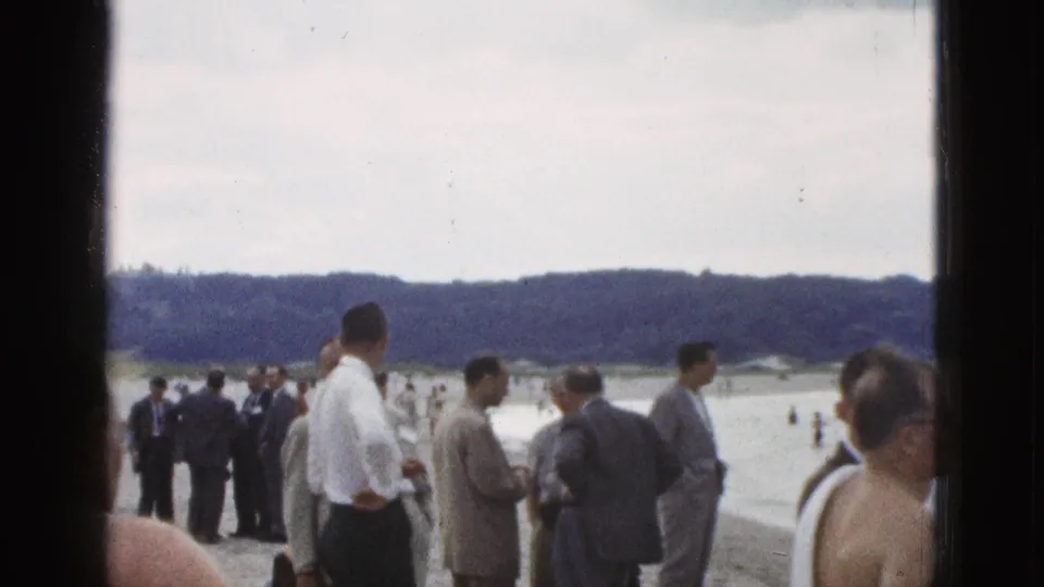1953 California shoreline view with suited men, casual beachgoers, a shirtless figure, and swimmers spread across the water behind them.