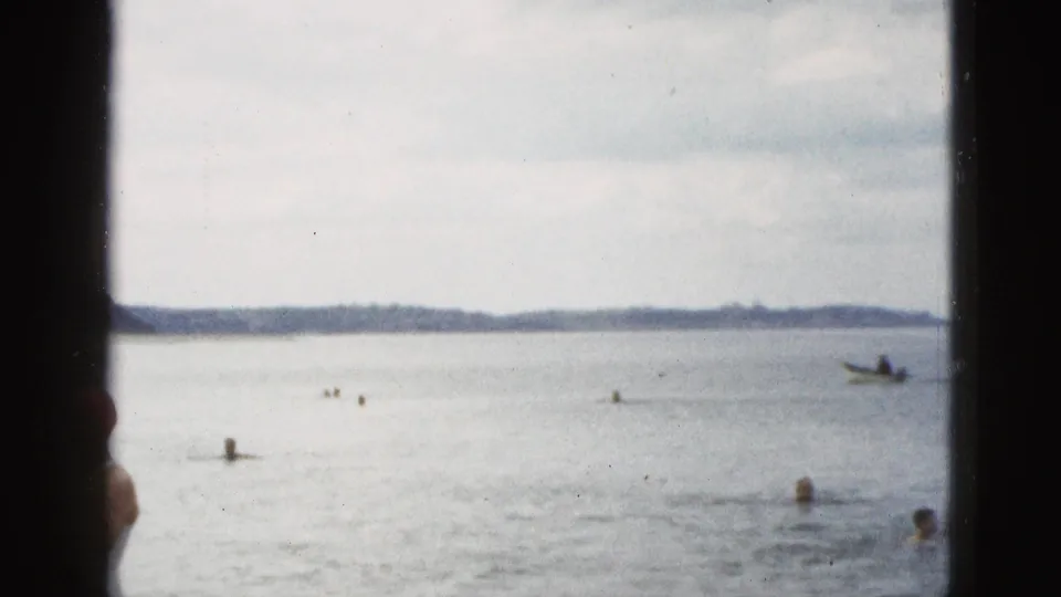 1953 California beach view looking out over swimmers in the water, a distant shoreline, and a small boat near the right side of the frame.