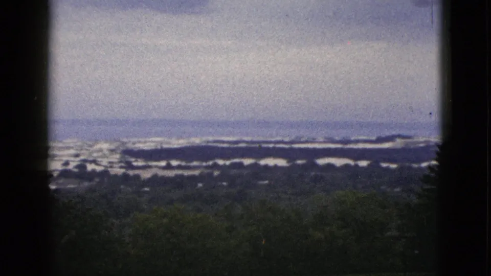 1953 California 8mm frame showing dark side edges, a dense green foreground, and distant water or snow-like bands beneath a pale sky.