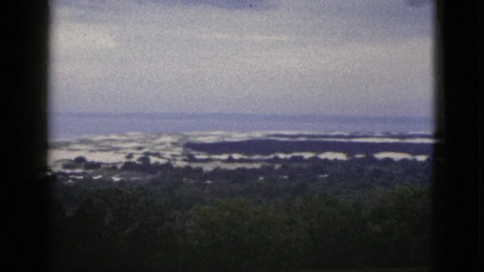 1953 California home-movie frame with dark framing at the sides, treetops below, and distant water or snow bands under muted sky.