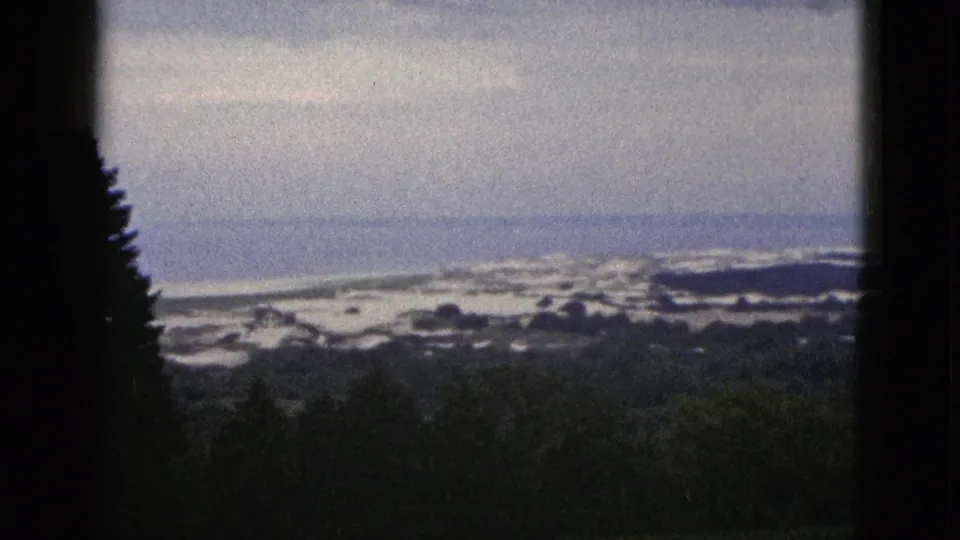 California 8mm frame from 1953 showing a quiet coastal panorama with dark trees, surf bands, and muted ocean light.
