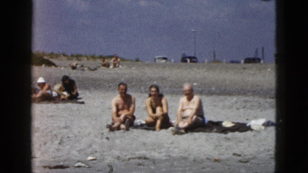 1953 California home-movie frame showing three adults seated on sand, with beachgoers and distant cars behind them.