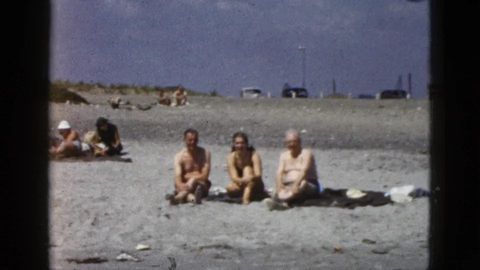 1953 California home-movie frame showing three adults seated on sand, with beachgoers and distant cars behind them.