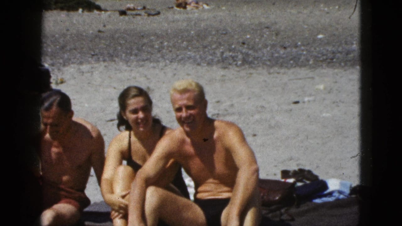 Three adults in swimsuits sit on sand at a beach, with two looking toward the camera.