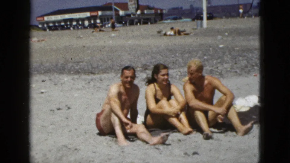 Three friends sit on sand at a California beach, with a beachfront structure, cars, and distant bathers behind them.