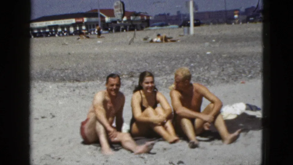 Three adults in swimsuits sit together on the sand at a 1953 California beach, smiling toward the camera.