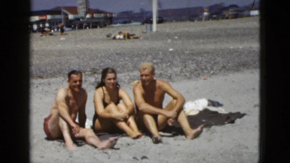 Three friends remain seated on a sunny California beach, with towels nearby and mid-century beachfront activity in the background.