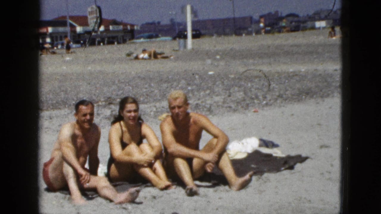 Three adults sit together on a California beach in 1953 home-movie footage, with distant buildings, cars, and sunbathers behind them.