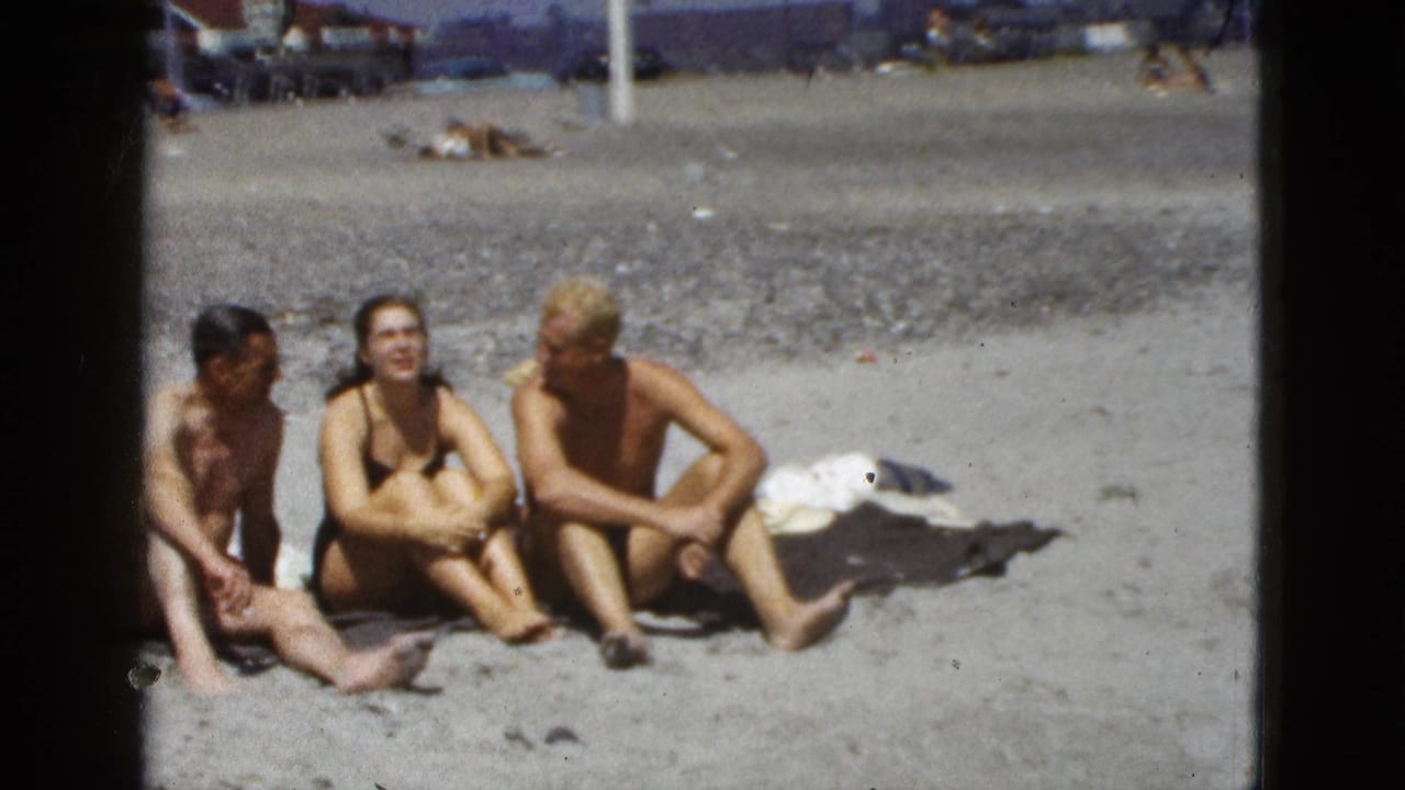 Three adults sit on beach sand near towels, with a distant beach area and buildings behind them.