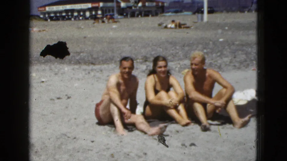A 1953 California beach home-movie frame shows three adults seated on sand, with background buildings and visible film wear.