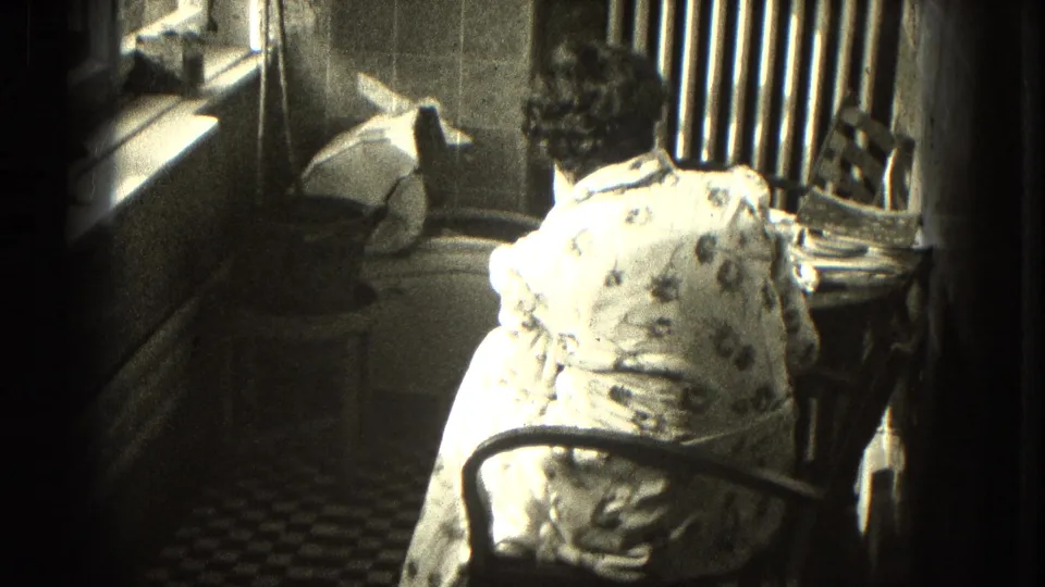 In a Milwaukee domestic interior, a woman in a patterned dress sits from behind at a writing desk near a radiator and sunlit window.