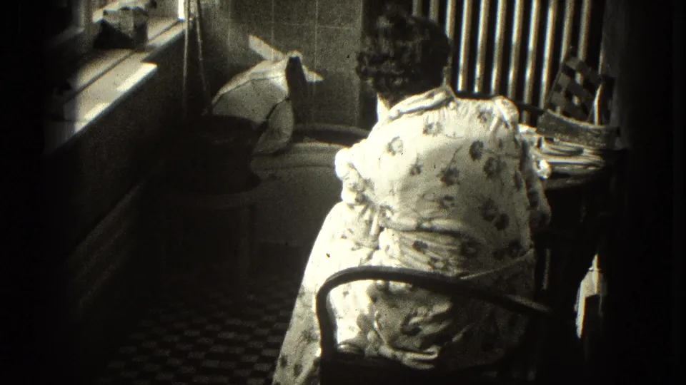 Closer 8mm view of the woman seated at a desk in the 1953 Milwaukee home interior, framed by window light and checkered flooring.