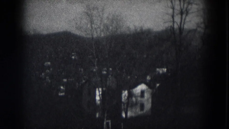 1953 Milwaukee home-movie frame showing distant suburban houses among leafless trees beneath a dark wooded ridge.