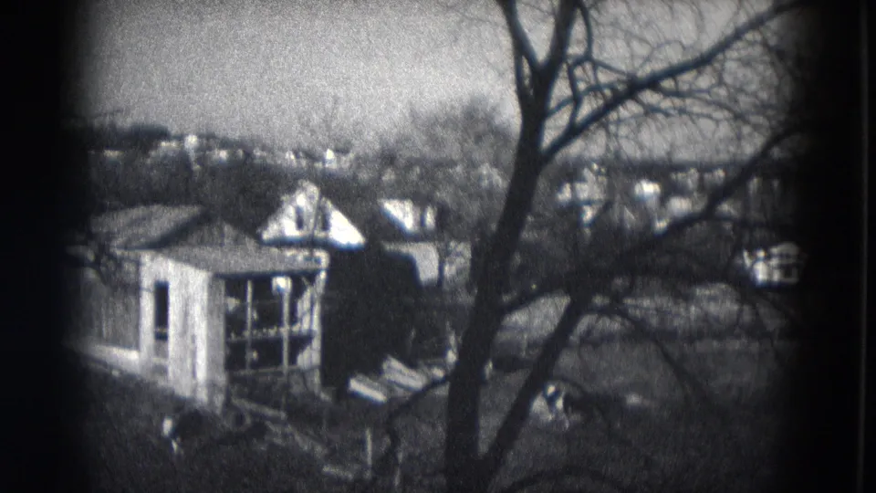 1953 Milwaukee 8mm frame showing a small residential structure, bare tree branches and distant rooftops beyond it.