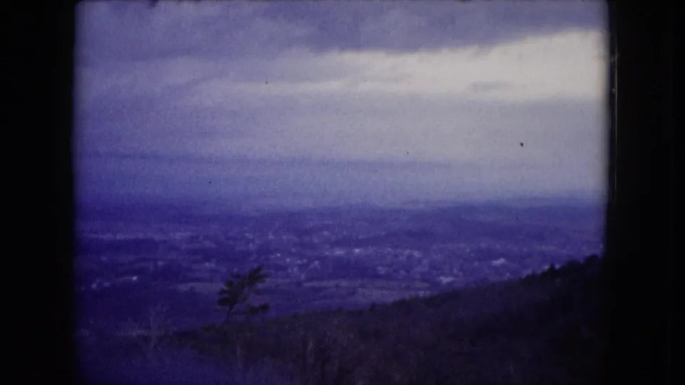 1953 Milwaukee-area home-movie frame showing a hazy distant city or shoreline beneath a cloudy sky, with dark hillside vegetation at the bottom.