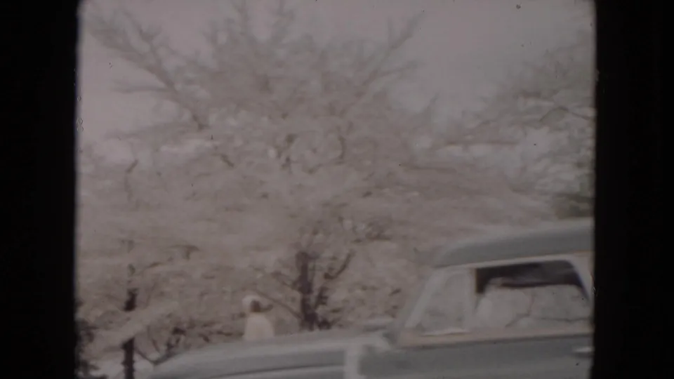 1953 winter home-movie frame showing a rounded classic car near snowy trees, with a partially visible person or passerby in the background.