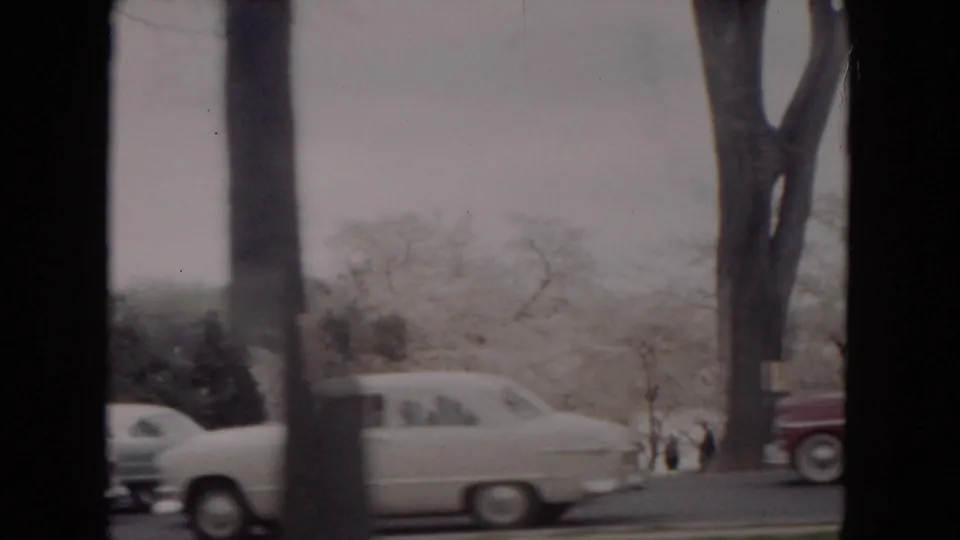 A white 1950s sedan passes a snowy Milwaukee roadside with bare trees and small distant pedestrians.
