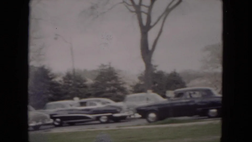 Parked 1950s automobiles line a snowy Milwaukee roadside, with bare trees, evergreens, and winter grass visible.