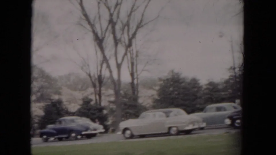 1953 Milwaukee home-movie frame showing parked rounded automobiles beside a bare, tree-lined road.