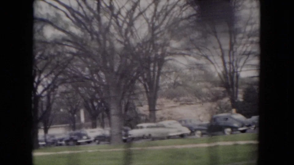 1953 Milwaukee frame with several parked cars beside grass and leafless trees, filmed from a passing 8mm viewpoint.