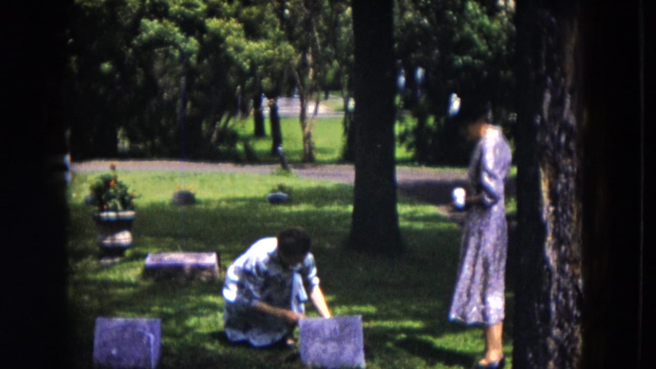 Two women in dresses stand and kneel among grave markers on a sunny cemetery lawn.