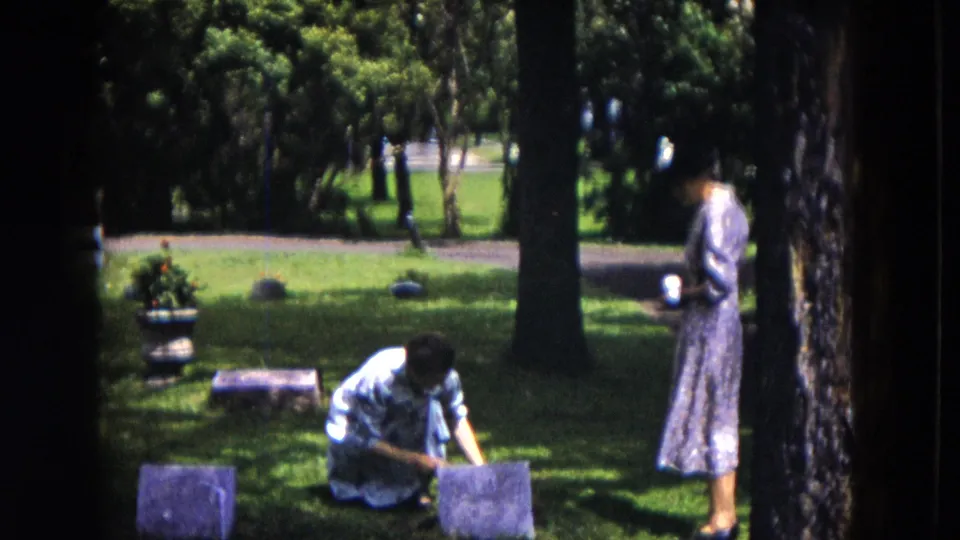 In a 1953 Milwaukee cemetery scene, one woman kneels near a grave marker while another stands beside a tree holding a cup.