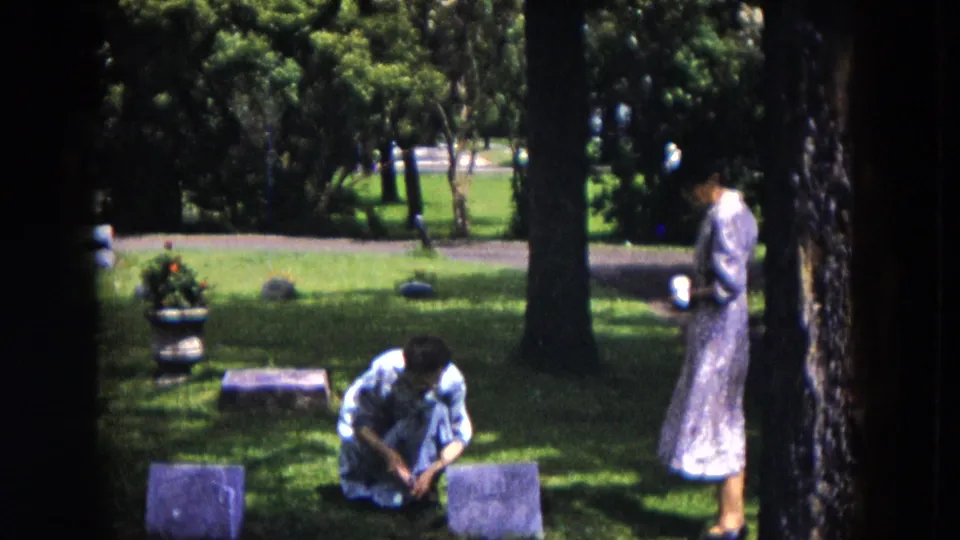 A woman kneels at a grave marker in a Milwaukee cemetery while a second woman in a patterned dress stands near a tree.