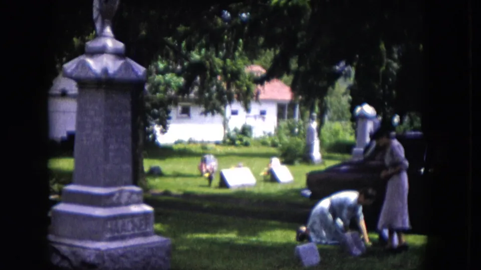 A tall monument anchors the foreground as two women tend smaller grave markers on a grassy Milwaukee cemetery lawn.