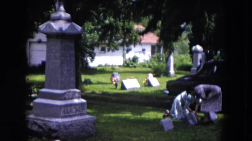Two women bend close to grave markers in a 1953 Milwaukee cemetery, with trees, a house, and a parked car in the background.