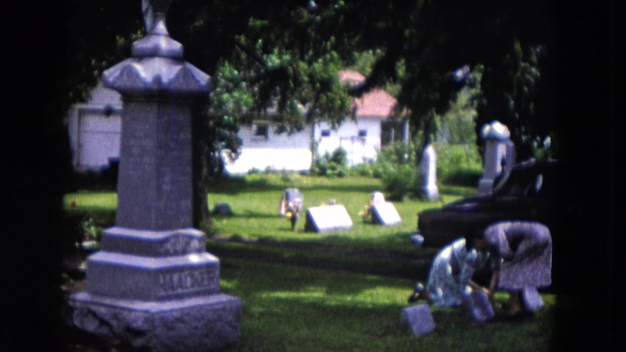 Two women bend near graves in a cemetery beside a large gray monument.
